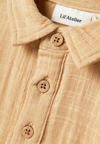 Beež kaelusega särk, mis on valmistatud tekstuuriga kangast, millel on nähtav ruuduline muster, funktsioneerides kahe puidust nupuga, millel on lillemustrid, ning kaubamärgi silt.