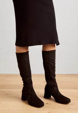 Bottes hautes jusqu'au genou en daim noir avec un bout carré et un talon carré, dotées d'une fermeture éclair latérale et d'une texture lisse. Portées avec une jupe noire à côtes.