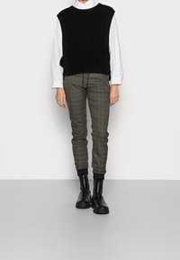 Person klädd i en vit långärmad skjorta, svart ärmlös stickad väst, grå rutig byxor och svarta ankelskor som står på ett ljust golv.