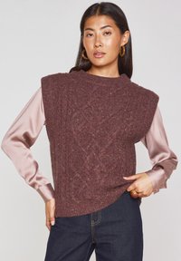 Gilet lavorato a maglia di un intenso bordeaux con un motivo a trecce testurizzato, senza maniche e con colletto a costine, indossato sopra una blusa di seta rosa chiaro.