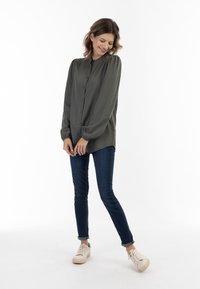 Donkergroen overhemd met lange mouwen, een knoopsluiting, opbollende schouders en manchetten, gecombineerd met donkere skinny jeans en lichtgekleurde sneakers.