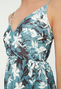 Blaues ärmelloses Kleid mit weißen und dunklen Blumenmuster, das über einer oberen Körperhälfte mit V-Ausschnitt und gerafftem Taillendetail getragen wird.