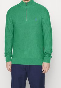 Grön stickad tröja med hög krage och halvdragkedja framtill. Har ribbade muddar och fåll. Litet blått logo på övre vänstra sidan.