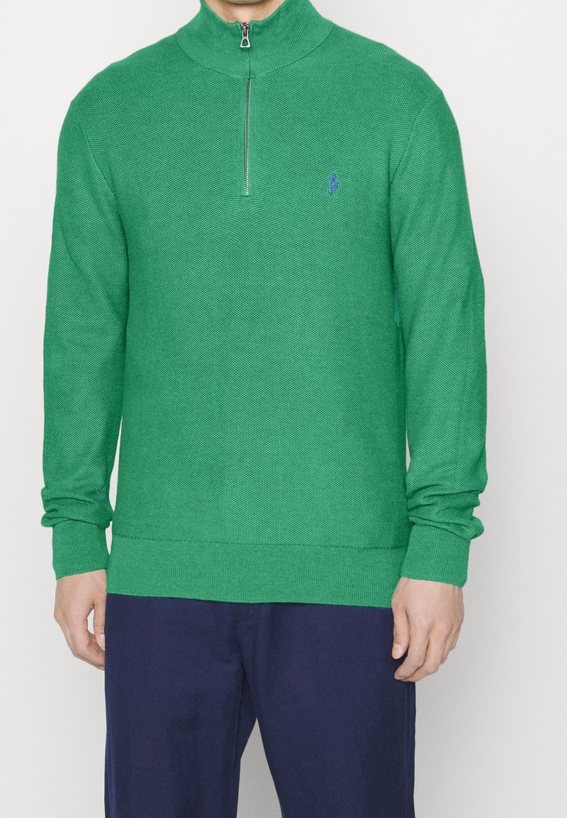 Grön stickad tröja med hög krage och halvdragkedja framtill. Har ribbade muddar och fåll. Litet blått logo på övre vänstra sidan.