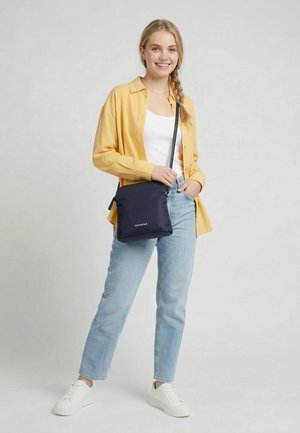 Junge Frau lächelt, trägt ein gelbes Hemd, weißen Top, hellblaue Jeans, weiße Turnschuhe und trägt eine schwarze Umhängetasche mit Logo.