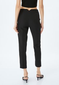 Pantalones negros a medida con pierna entallada, que presentan un diseño en forma de V en la parte trasera, costuras verticales y una textura suave, combinados con sandalias de tacón negras.