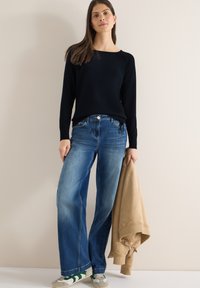 Jeune femme debout contre un mur clair, portant un pull noir, un jean large bleu, des baskets blanches, et tenant une veste beige.