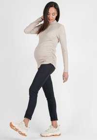 Maternity top in beige ribbed fabric with a mock neck, featuring side ruching. Paired with fitted black pants and white sneakers with orange accents.