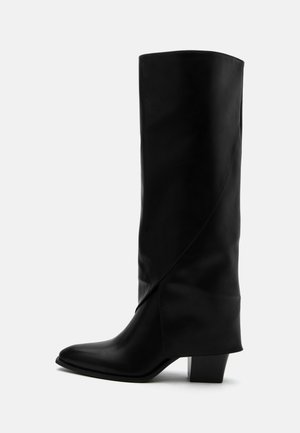 Botte haute en cuir noir avec couture inclinée, bout pointu et talon épais de taille moyenne sur fond blanc.