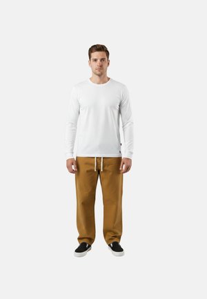 Uomo in piedi, di fronte, che indossa una camicia bianca a maniche lunghe, pantaloni marroni con coulisse e scarpe nere slip-on su uno sfondo bianco uniforme.