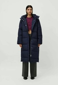 Mulher vestindo um longo casaco acolchoado azul-marinho sobre um top roxo cropped com botões, calças largas cinza e sapatos pretos, em pé contra um fundo simples.