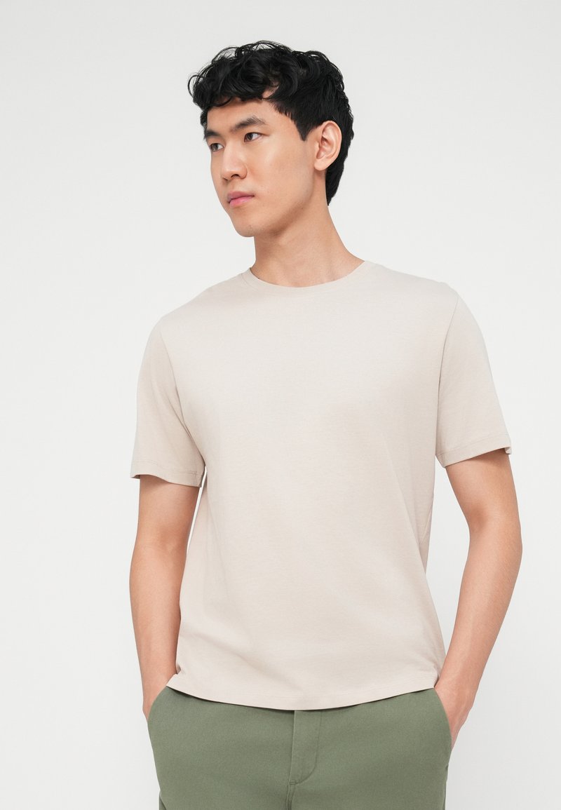 Joven con cabello negro, vistiendo una camisa beige de manga corta y pantalones verdes, de pie con las manos en los bolsillos, mirando hacia la izquierda.