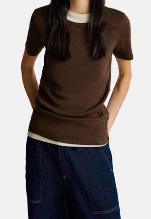 Personne portant une chemise marron à manches courtes par-dessus une chemise beige clair avec un pantalon en denim bleu, les mains dans les poches du pantalon.