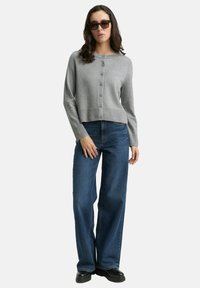 Graue, kurze Strickjacke mit Rundhalsausschnitt und zentraler Knopfleiste, kombiniert mit blauen weiten Jeans und schwarzem Schuhwerk.