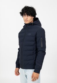 Navy Padded Jacke mit Reißverschluss vorne, Kapuze und Rippbündchen. Verfügt über eine Reißverschlusstasche am Ärmel und ein Logo auf der Brust.