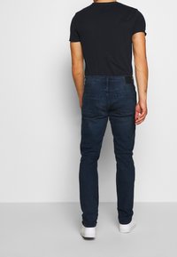 Tmavě modré džíny se střihm slim fit, s hladkou texturou, pěti kapsami a koženou nášivkou na zadním pásku. Kombinovány s černým tričkem.