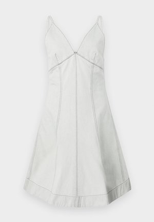 Femme tatouée portant une robe grise claire sans manches et des bottes noires mi-mollet, tenant un petit sac à main noir sur un fond uni.