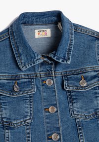 Blauwe spijkerjas voor kinderen met een kraag, metalen knopen met hartgraveer, twee borstzakken en een maatlabel dat 6-7 jaar aangeeft.