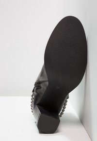 Chaussure à talon haut en cuir noir avec un bout carré, une semelle texturée, un détail de chaîne sur le côté, et la taille indiquée à l'intérieur comme 37.