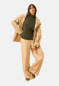 Giacca impermeabile beige sopra un maglione verde scuro, abbinata a pantaloni larghi beige chiaro e sneakers marroni. Il tessuto appare liscio e informale.