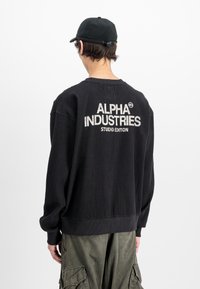 Person wearing black textured sweatshirt with "Alpha Industries Studio Edition" text on back and green cargo pants, facing away.