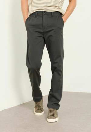 Mann trägt schwarze Hose mit geradem Bein, hellbraune Wildledersneaker mit weißen Sohlen und ein beiges gestreiftes Hemd, steht mit den Händen in den Taschen.