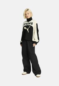Black and cream zip-up jacket with large "PUMA" logo and cat graphic. Loose black pants. White sneakers with tan soles.