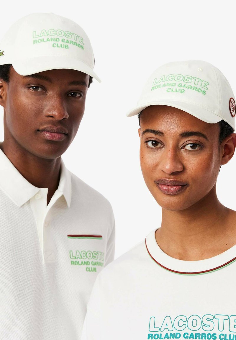 Gorras blancas con el texto bordado en verde "LACOSTE ROLAND GARROS CLUB"; el hombre lleva una polo con un detalle de bolsillo a rayas rojas y verdes.