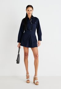 Robe chemise en coton bleu marine avec manches longues, devant boutonné et jupe plissée. Associée à des sandales à plateforme et un petit sac à main.