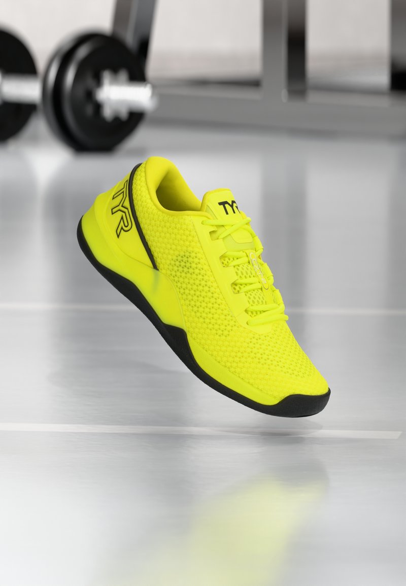 Neongele sport schoen met mesh textuur, zwarte accenten en een gevoerde tong. Heeft een gebogen vorm en een vetersluiting.