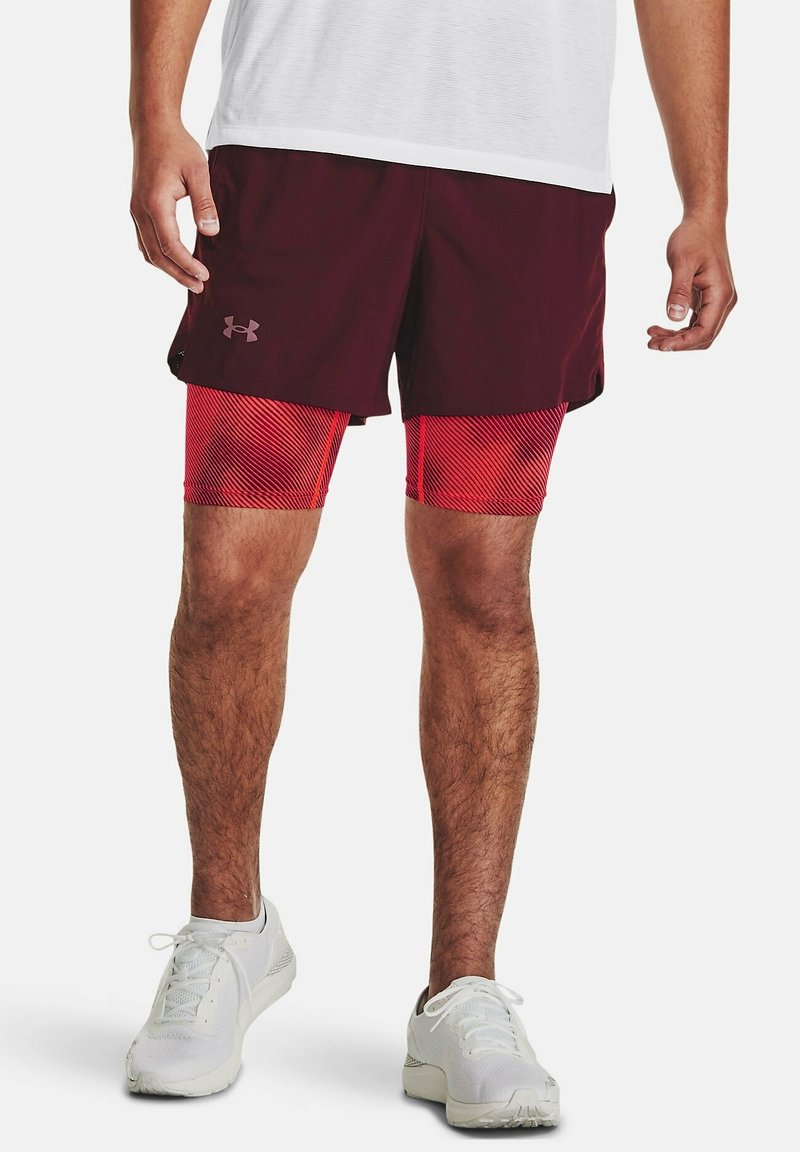 Homme portant un short bordeaux par-dessus des leggings de compression rouges et des chaussures de sport blanches, debout devant un fond blanc.