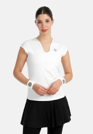 Femme portant un haut blanc à manches courtes avec logo, une jupe noire, et des bracelets blancs assortis avec logo, se tenant les mains croisées.