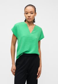 Γυναίκα με πλεξούδες που φοράει μια υφασμάτινη πράσινη μπλούζα με κοντά μανίκια και μαύρο παντελόνι, στέκεται μπροστά σε ένα απλό λευκό φόντο.