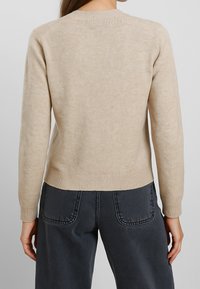 Maglione lavorato a maglia beige con maniche lunghe, polsini e orlo a coste, dotato di scollatura rotonda e una texture liscia e morbida.