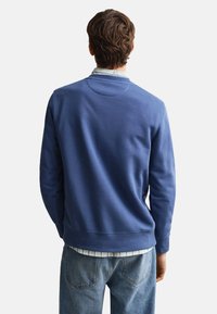 Man with brown hair wearing a blue sweatshirt over a striped shirt and blue jeans, facing away against a plain white background.