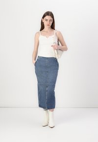 Bílý bezrukávový top s výstřihem do V, zkombinovaný s modrou džínovou maxi sukni. Modelka nosí bílé kotníkové boty a drží malou bílou tašku.