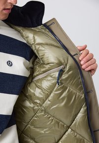 Puffersjakke i olivengrøn med en lynlåslomme indeni, glat quiltet tekstur og krave i marineblå corduroy.