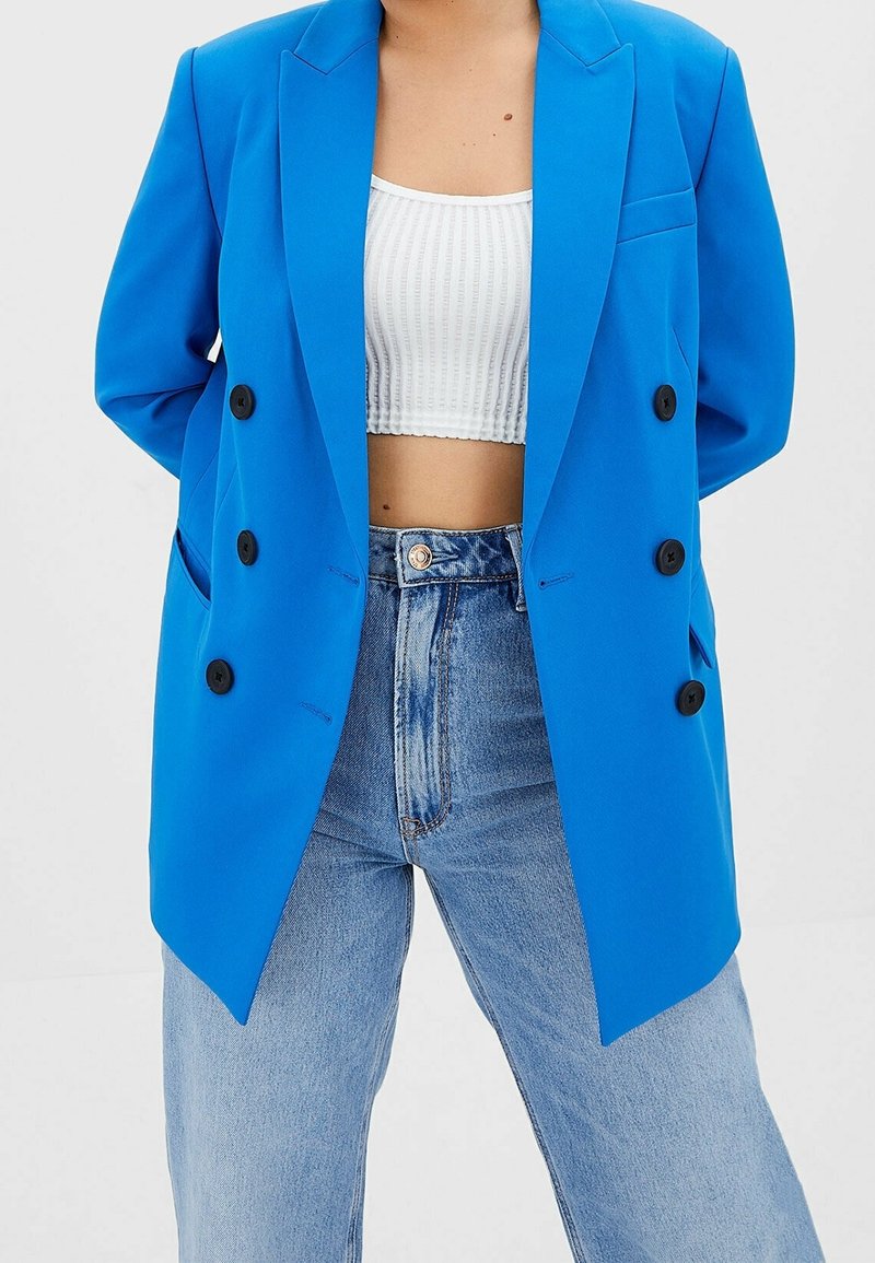 Blazer bleu croisé à boutons noirs, épaules structurées et poches latérales, associé à un crop top blanc et un jean en denim clair.