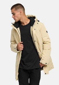 Beige isolerad jacka med huva, med svart foder, tryckknappar och fickor med dragkedja. Stylad över en svart t-shirt.