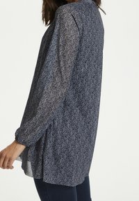 Vrouw in een langemouwen marineblauwe tuniek met een klein wit loofpatroon, gecombineerd met donkerblauwe jeans, staande tegen een effen achtergrond.