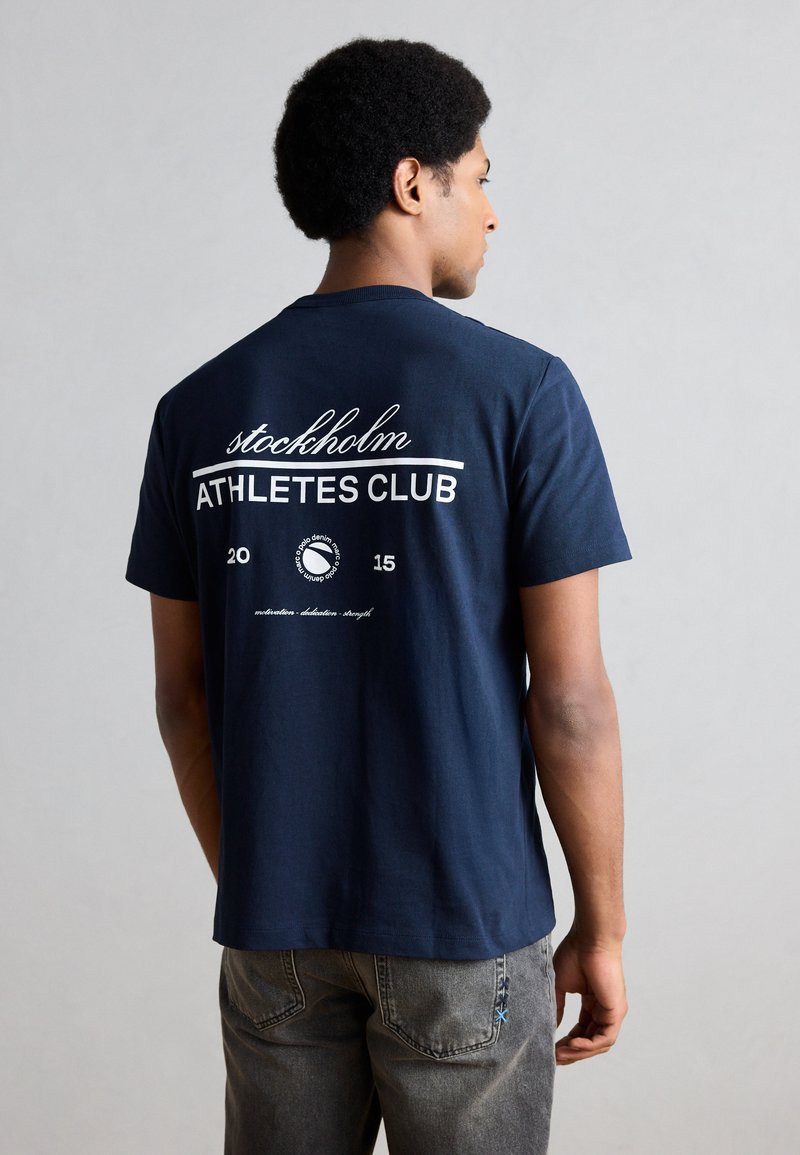 Sötétkék pamutpóló, hátul fehér felirattal: "stockholm ATHLETES CLUB", további részletekkel alatta. Egyszerű kerek nyakú kialakítás.