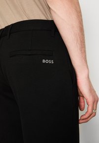 Gros plan sur le côté d'une personne portant un pantalon noir avec un logo "BOSS" près de la poche arrière et une chemise beige rentrée.