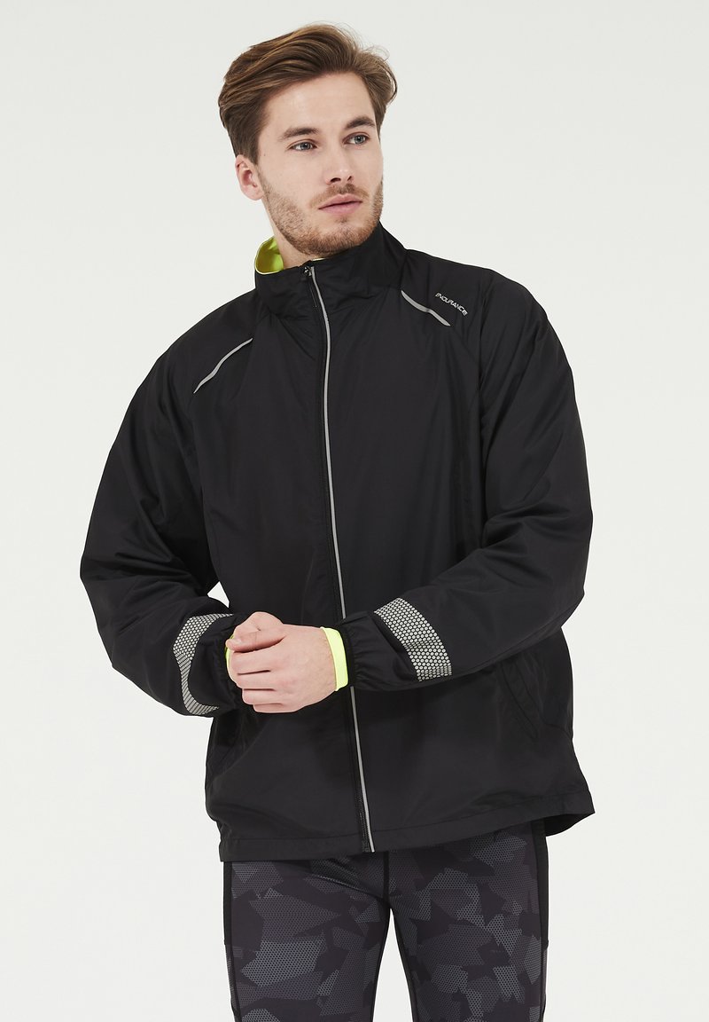 Veste de sport noire avec un col montant, des accents réfléchissants et des détails en mesh sur les manches, associée à des leggings à motifs.