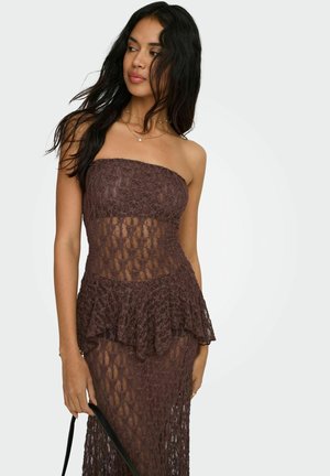 Kvinde med langt mørkt hår iført en brun stropløs blondekjole, som holder en sort håndtaske og kigger let til siden.