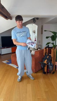 Νέος άνδρας με ανοιχτό μπλε πουκάμισο Jack & Jones και γκρι φόρμα, στέκεται σε ένα σαλόνι κρατώντας διπλωμένα ρούχα κοντά σε ένα καφέ καναπέ και μηχάνημα γυμναστικής.