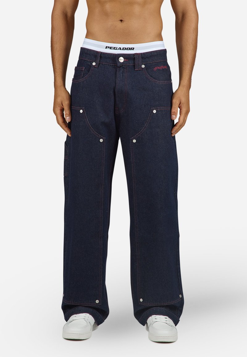 Dark blue denim trousers with a wide leg cut, featuring red stitching, multiple pockets, and metal snap accents. Includes a white waistband.