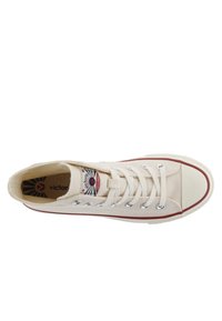 Canvas sneakers in crème met rode accenten, ronde witte neus, vetersluiting aan de voorkant en een logo patch nabij de tong. Gladde textuur.