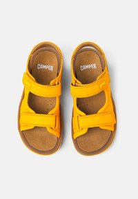 Keltaiset sandaalit, joissa on kaksi säädettävää tarranauhaa, pehmeä ruskea sisäpohja ja kestävä pohja. Brändin nimi "CAMPER" näkyy pohjassa.