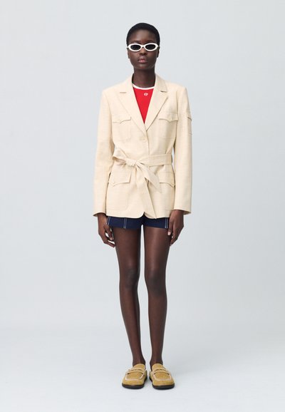Blazer beige clair avec une ceinture et deux poches avant, porté sur une chemise rouge et un short noir. Des lunettes de soleil blanches et des mocassins jaunes complètent le look.