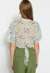 Blusa in chiffon floreale con maniche corte a volant, vita stretta con un nastro e chiusura a bottone sul retro. I colori includono rosa pastello e viola.